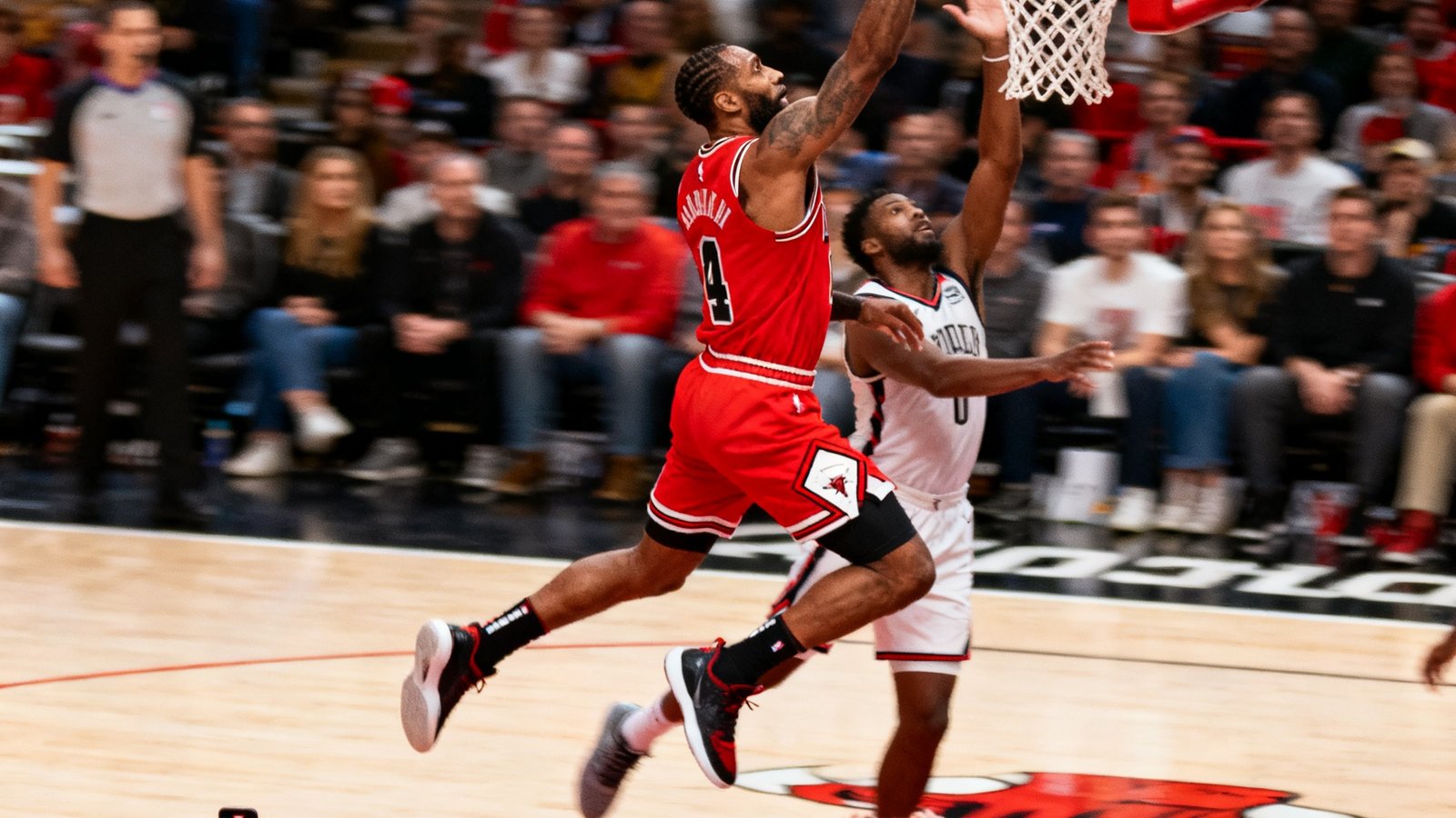 NBA赛场上一位身穿红色球衣的球员在快攻中腾空而起，单臂暴扣的瞬间，防守球员试图封盖但已来不及，篮筐周围的观众纷纷举起手机拍摄
